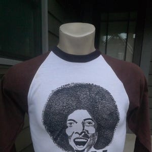 May include: A white t-shirt with brown sleeves features a black and white illustration of a man with an afro and the word "SLY" printed below.