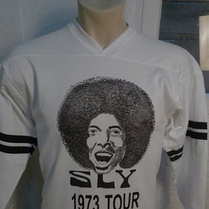May include: White long-sleeved shirt with black stripes on the sleeves. The shirt features a black and white illustration of Sly Stone with the text "SLY 1973 TOUR".