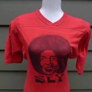 May include: Red t-shirt with a black and white image of Sly Stone, the lead singer of Sly and the Family Stone, with the word "SLY" printed below the image.