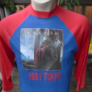 Pode incluir: Uma camiseta de estilo beisebol azul e vermelha com um logotipo de banda e o texto "1981 TOUR".