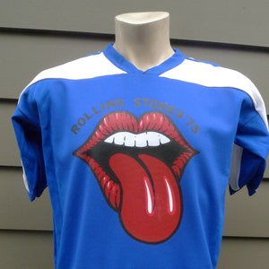 May include: A blue short-sleeved t-shirt with white trim on the collar and sleeves. The shirt features a red and white graphic of the Rolling Stones tongue logo with the text "Rolling Stones '75" above it.