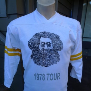 May include: White long-sleeved t-shirt with yellow stripes on the sleeves. The shirt features a black and white illustration of a man with long hair and a beard. The text "1978 TOUR" is printed below the illustration.