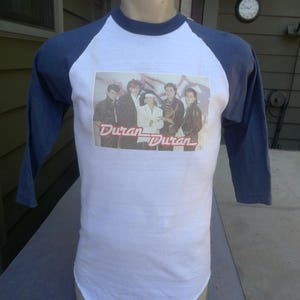 May include: White and navy blue baseball style t-shirt featuring a band photo and the text "Duran Duran" in red. The shirt has three-quarter length sleeves and is displayed on a mannequin.