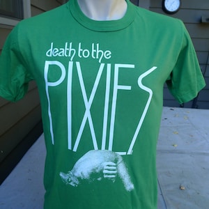 May include: A bright green short-sleeve t-shirt featuring the white text "death to the PIXIES" printed across the chest. Below the text is a white graphic illustration of a creature in a curled position on the lower front of the shirt.