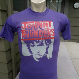 Puede incluir: Una camiseta morada de manga corta con un estampado gráfico del rostro de una persona en gris claro y el texto JOHNNY THUNDERS impreso en letras rojas negritas en la parte superior. La camiseta tiene cuello redondo y corte estándar.