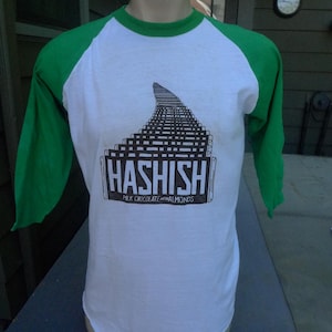 May include: White and green raglan-style t-shirt with a graphic that reads "HASHISH" in a rectangular box. The graphic features a black and white design. The text "MILK CHOCOLATE with ALMONDS" is below the word "HASHISH".
