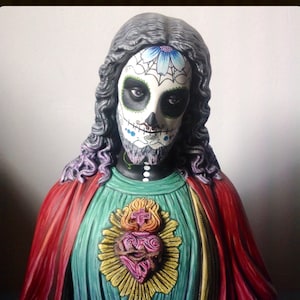 Może przedstawiać: Popiersie postaci z twarzą czaszki cukrowej z Dnia Zmarłych, długimi ciemnymi włosami i czerwonym płaszczem. Postać nosi turkusową tunikę ze złotym emblematem w kształcie serca. Podstawa jest ciemnoszara z napisem "MUERTIFY" i "2013".