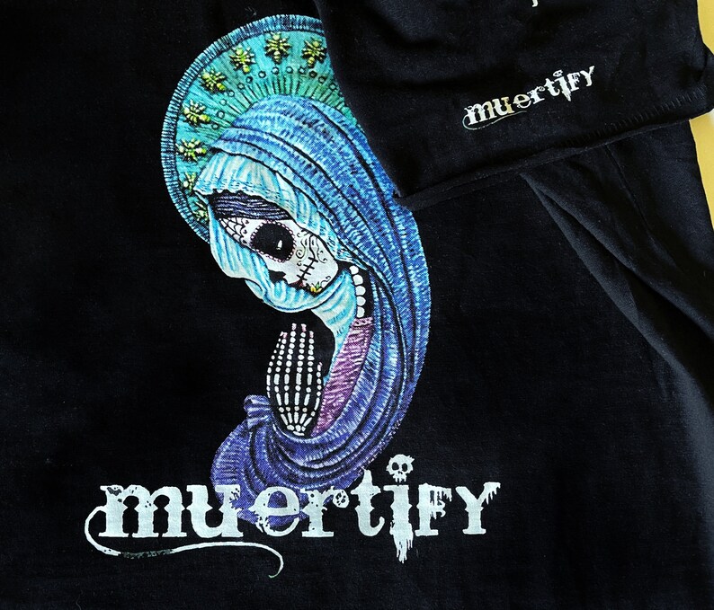 Puede incluir: Camiseta negra con una ilustraci&oacute;n azul y morada de una mujer con cara de calavera y la palabra "muertify" impresa en blanco.