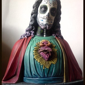 Może przedstawiać: Rzeźba popiersia przedstawiająca postać z pomalowaną twarzą czaszki, długimi ciemnymi włosami i czerwoną i turkusową szatą. Postać ma dekoracyjne serce na piersi. Widoczne są teksty "Muertify 2013" i "Muertefy".