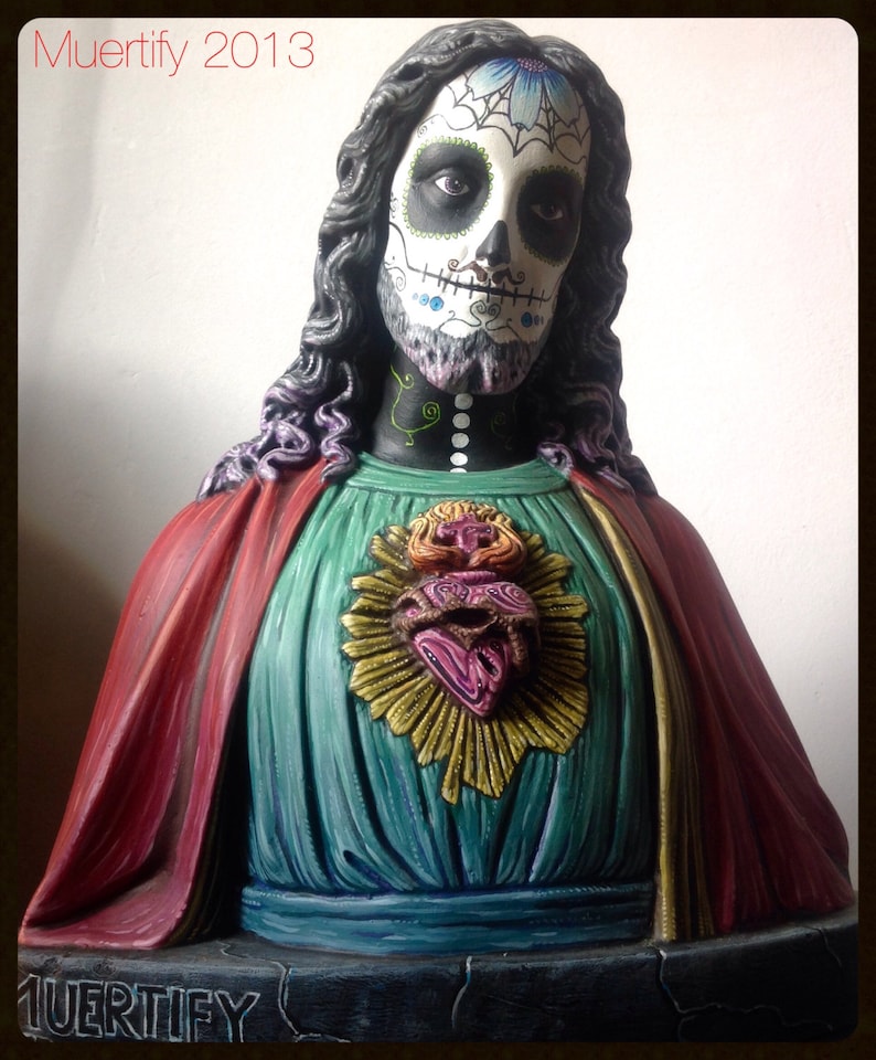 Może przedstawiać: Rzeźba popiersia przedstawiająca postać z pomalowaną twarzą czaszki, długimi ciemnymi włosami i czerwoną i turkusową szatą. Postać ma dekoracyjne serce na piersi. Widoczne są teksty "Muertify 2013" i "Muertefy".