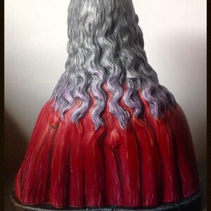 Może przedstawiać: Rzeźba przedstawiająca plecy postaci z długimi, falistymi, szarymi włosami z fioletowymi pasemkami. Postać ma na sobie żywą czerwoną szatę. Podstawa jest ciemna i popękana. Tekst "Muertify 2013" jest widoczny na dole.