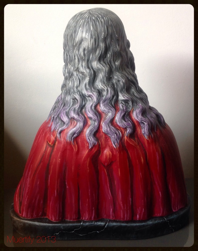 Może przedstawiać: Rzeźba przedstawiająca plecy postaci z długimi, falistymi, szarymi włosami z fioletowymi pasemkami. Postać ma na sobie żywą czerwoną szatę. Podstawa jest ciemna i popękana. Tekst "Muertify 2013" jest widoczny na dole.