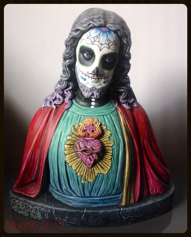 Może przedstawiać: Popiersie postaci z twarzą czaszki cukrowej z Dnia Zmarłych, długimi ciemnymi włosami i czerwonym płaszczem. Postać nosi turkusową tunikę ze złotym emblematem w kształcie serca. Podstawa jest ciemnoszara z napisem "MUERTIFY" i "2013".