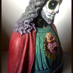 Może przedstawiać: Dekoracyjne popiersie z motywem Dnia Zmarłych. Postać ma pomalowaną twarz czaszki ze skomplikowanymi detalami, długie szare włosy oraz czerwoną i zieloną szatę. Podstawa jest ciemnoszara z napisem "MUERTIFY" i "2013".