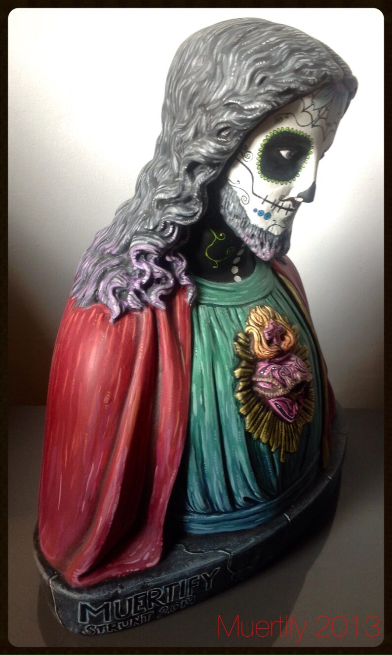 Może przedstawiać: Dekoracyjne popiersie z motywem Dnia Zmarłych. Postać ma pomalowaną twarz czaszki ze skomplikowanymi detalami, długie szare włosy oraz czerwoną i zieloną szatę. Podstawa jest ciemnoszara z napisem "MUERTIFY" i "2013".