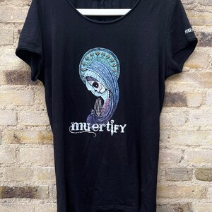 Puede incluir: Camiseta negra con un estampado gr&aacute;fico de una figura esquel&eacute;tica que lleva una t&uacute;nica azul y un halo. El texto "muertify" est&aacute; impreso debajo de la figura.