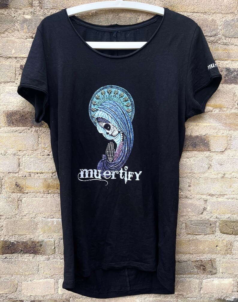 Puede incluir: Camiseta negra con un estampado gr&aacute;fico de una figura esquel&eacute;tica que lleva una t&uacute;nica azul y un halo. El texto "muertify" est&aacute; impreso debajo de la figura.