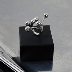 Op de afbeelding: Een zilverkleurige ring met een cluster van bolvormige vormen. De ring wordt gepresenteerd op een kleine zwarte doos tegen een metallic achtergrond. Het ontwerp bevat meerdere bollen van verschillende groottes.