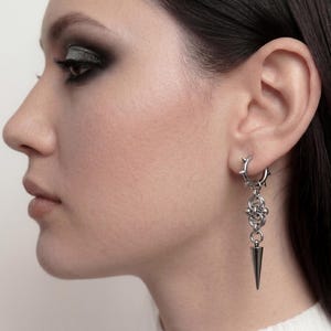 Könnte beinhalten: Silberfarbene Ohrringe. Ein Ohrring ist ein kleiner Reifen mit dornenartigen Details. Der andere hat ein Kettenmuster mit einem hängenden, spitz zulaufenden Kegel. Die Ohrringe werden am Ohr getragen.