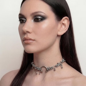 Könnte beinhalten: Ein silberfarbenes Choker-Halsband mit aufwendigen floralen und baumelnden Perlen Details. Die Halskette hat ein zentrales kreisförmiges Design und wird um den Hals getragen. Der Schmuck wird vor einem neutralen Hintergrund präsentiert.