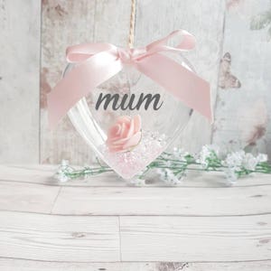 Puede incluir: Un adorno de corazón de vidrio transparente con una cinta rosa y una rosa rosa en su interior. La palabra "mum" está escrita en la parte delantera del adorno.