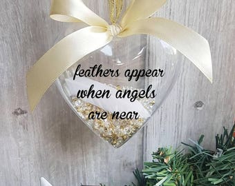 May include: A clear heart-shaped ornament with gold glitter and a white feather inside. The ornament has a white ribbon and a gold string for hanging. The ornament has the text "feathers appear when angels are near" printed on it.