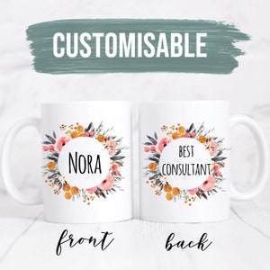 May include: White ceramic mug with a floral wreath design. The front of the mug says "Nora" in a circle. The back of the mug says "Best Consultant" in a circle.