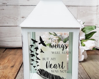Puede incluir: Linterna blanca con un panel de vidrio que presenta una pluma negra y el texto "Tus alas estaban listas pero mi corazón no lo estaba". El panel de vidrio también presenta una silueta de pájaros volando.