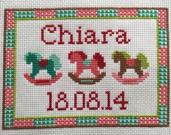 May include: Cross-stitch embroidery with a pink, green, and brown border. The center features the name Chiara, three rocking horses, and the date 18.08.14.