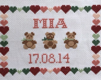 May include: Cross-stitch embroidery with a pink heart border, featuring the name "MIA" in pink lettering, three brown teddy bears, and the date "17.08.14" in pink lettering.