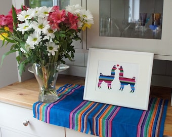 May include: A white picture frame with a colorful cross-stitch design of two llamas kissing. The frame is sitting on a blue and multi-colored striped table runner.