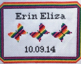 May include: A white cross-stitch embroidery with a rainbow border. The embroidery features the name "Erin Eliza" and the date "10.09.14" with three rainbow butterflies.
