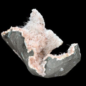 Pode incluir: Amostra mineral com cristais rosa pálido e formações brancas em forma de agulha. Os cristais estão inseridos numa matriz cinza escuro, com bordas verdes e brancas. O fundo é preto.
