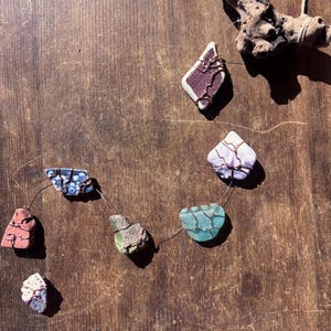 May include: A handmade necklace featuring sea glass pieces in various colors, including red, blue, green, and purple, each wrapped with copper wire. The necklace is displayed on a wooden surface with a piece of driftwood.