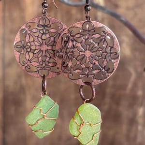 May include: Copper earrings featuring a circular, openwork design with a floral pattern. Each earring has a green sea glass pendant wrapped in copper wire. The earrings hang from copper-colored ear wires, creating a unique, handcrafted look.