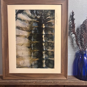 May include: Abstract art print in a wooden frame. The artwork features a dark, textured central design with lighter, mottled areas. The frame is a warm brown, and the print is signed.