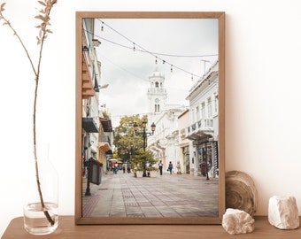 Op de afbeelding: Een ingelijste foto van een straatbeeld in een stad met witte gebouwen, een klokkentoren en lichtjes boven de straat. Mensen lopen over straat.