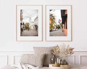 Op de afbeelding: Twee ingelijste foto's van een straatbeeld in een stad. De linker foto toont een straat met gebouwen aan beide kanten en mensen die lopen. De rechter foto toont een straat met een palmboom en een vlag die aan een gebouw hangt.