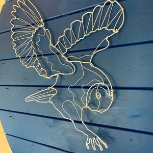 Pode incluir: Escultura de arame branco de um p&aacute;ssaro em voo, montada sobre um fundo de madeira azul. As asas do p&aacute;ssaro est&atilde;o abertas e seu corpo &eacute; detalhado com arame. Uma pe&ccedil;a decorativa para decora&ccedil;&atilde;o de parede.