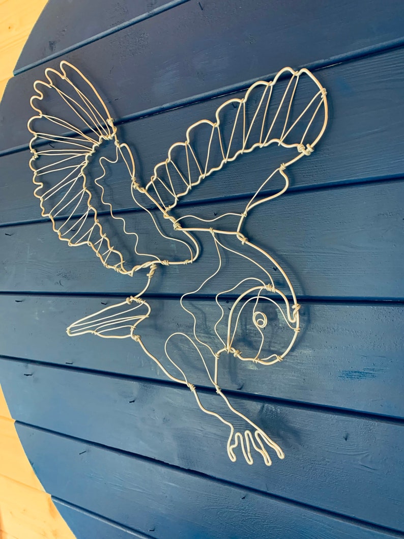 Pode incluir: Escultura de arame branco de um p&aacute;ssaro em voo, montada sobre um fundo de madeira azul. As asas do p&aacute;ssaro est&atilde;o abertas e seu corpo &eacute; detalhado com arame. Uma pe&ccedil;a decorativa para decora&ccedil;&atilde;o de parede.