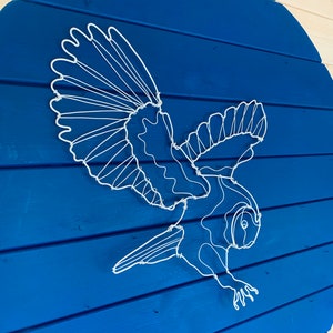 Pode incluir: Escultura de arame branco de uma coruja em voo, montada sobre um fundo redondo de madeira azul. As asas da coruja est&atilde;o abertas e seu corpo &eacute; detalhado com arame. O fundo &eacute; feito de t&aacute;buas de madeira horizontais.