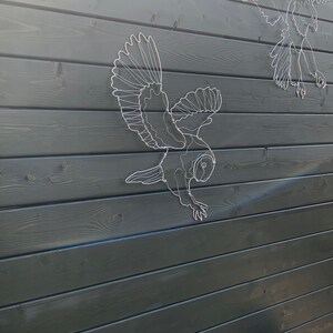 Pode incluir: Esculturas de arame branco de p&aacute;ssaros, incluindo uma coruja, montadas em uma cerca de madeira cinza escura. A escultura da coruja em primeiro plano est&aacute; em pleno voo com as asas abertas. O fundo mostra a textura das t&aacute;buas de madeira.