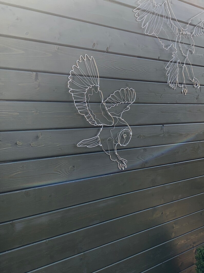 Pode incluir: Esculturas de arame branco de p&aacute;ssaros, incluindo uma coruja, montadas em uma cerca de madeira cinza escura. A escultura da coruja em primeiro plano est&aacute; em pleno voo com as asas abertas. O fundo mostra a textura das t&aacute;buas de madeira.
