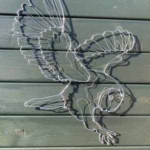 Pode incluir: Escultura artesanal em arame de metal de uma coruja em voo. A obra de arte &eacute; feita de arame prateado e &eacute; montada em um fundo de madeira verde. As asas da coruja est&atilde;o abertas e suas garras est&atilde;o estendidas.