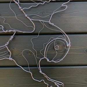 Pode incluir: Escultura artesanal em arame prateado representando um p&aacute;ssaro em voo. A obra est&aacute; montada sobre um painel de madeira verde escuro. A forma do p&aacute;ssaro &eacute; definida pelas curvas e dobras do arame, criando um contorno delicado.