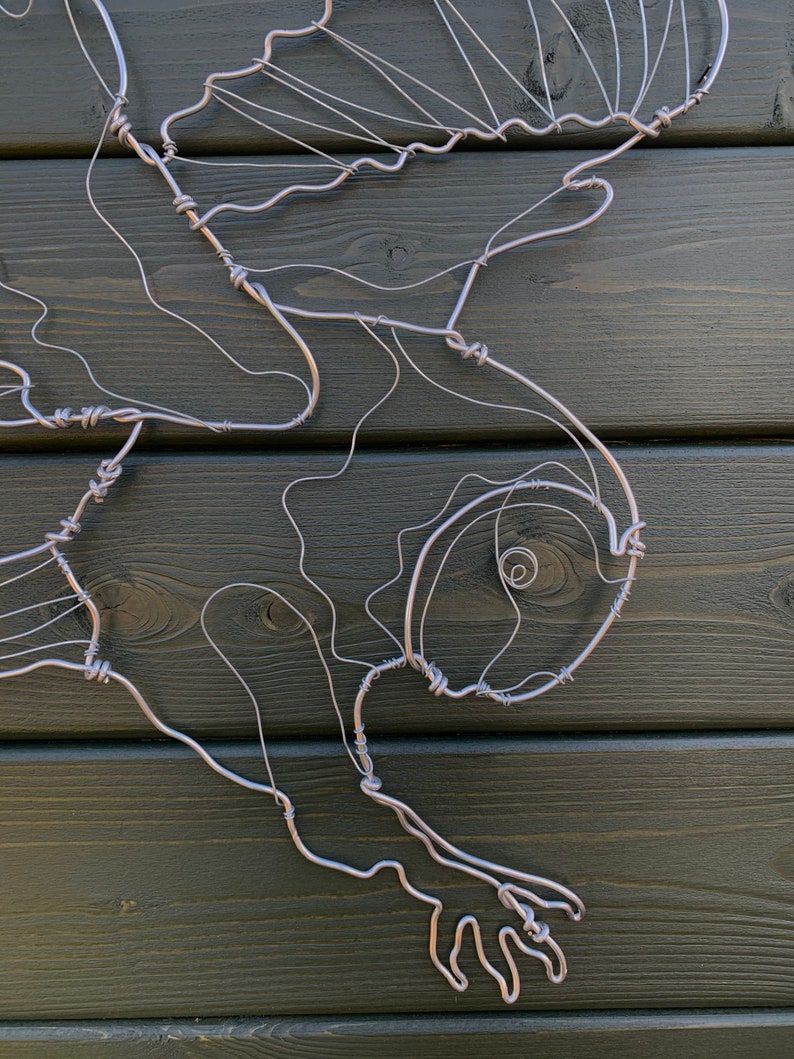 Pode incluir: Escultura artesanal em arame prateado representando um p&aacute;ssaro em voo. A obra est&aacute; montada sobre um painel de madeira verde escuro. A forma do p&aacute;ssaro &eacute; definida pelas curvas e dobras do arame, criando um contorno delicado.