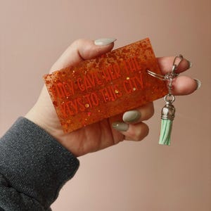 Op de afbeelding: Oranje rechthoekige sleutelhanger met glitter en de tekst "They Gave Her The Keys To This City". De sleutelhanger heeft een zilveren sluiting en een groene kwast. De achtergrond is effen roze.