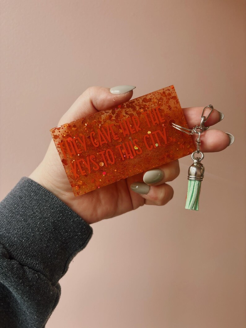 Op de afbeelding: Oranje rechthoekige sleutelhanger met glitter en de tekst "They Gave Her The Keys To This City". De sleutelhanger heeft een zilveren sluiting en een groene kwast. De achtergrond is effen roze.