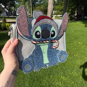 Pu&ograve; includere: Una borsa a tracolla grigio chiaro con un design scintillante del personaggio Stitch con un cappello da Babbo Natale e un bastoncino di zucchero. La borsa ha un bordo azzurro e dettagli a fiocco di neve, perfetta per i fan Disney.