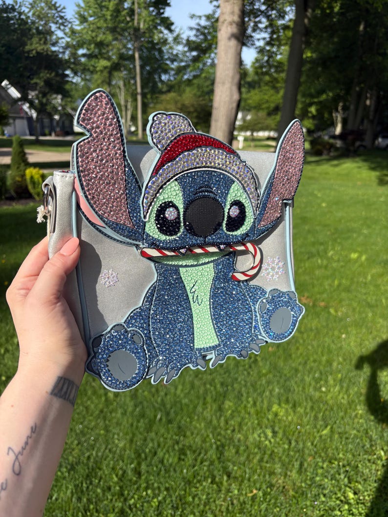 Pu&ograve; includere: Una borsa a tracolla grigio chiaro con un design scintillante del personaggio Stitch con un cappello da Babbo Natale e un bastoncino di zucchero. La borsa ha un bordo azzurro e dettagli a fiocco di neve, perfetta per i fan Disney.